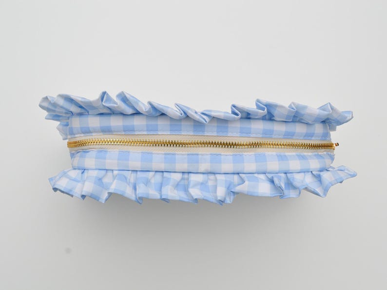 May include: A light blue and white gingham pattern cosmetic bag with a gold zipper. The bag is trimmed with ruffled fabric along the top and bottom edges. The bag is rectangular in shape.