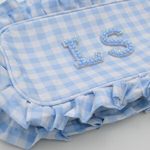 May include: A light blue and white gingham patterned pillow with a ruffled edge. The pillow features the letters "L" and "S" embellished with light blue rhinestones. The pillow is a decorative item.