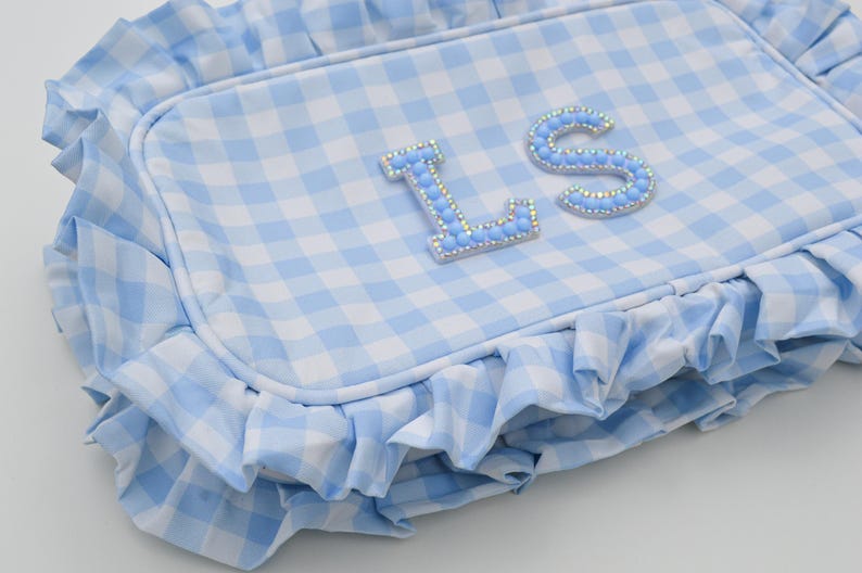 May include: A light blue and white gingham patterned pillow with a ruffled edge. The pillow features the letters "L" and "S" embellished with light blue rhinestones. The pillow is a decorative item.