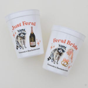 May include: Two white plastic cups with raccoon illustrations and text. One cup reads "Just Feral" with a champagne bottle graphic. The other cup says "Feral Bride" with a bouquet and rings. Both cups have "Amanda's Bachelorette" printed on them.