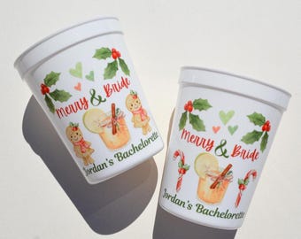 以下が含まれることがあります： お祝いのデザインが施された白いプラスチックカップ2つ。各カップには「Merry & Bride」と「Jordan's Bachelorette」の文字がプリントされています。ジンジャーブレッドマン、ヒイラギ、ハート、カクテルのイラストが特徴です。独身最後のパーティーに最適です。
