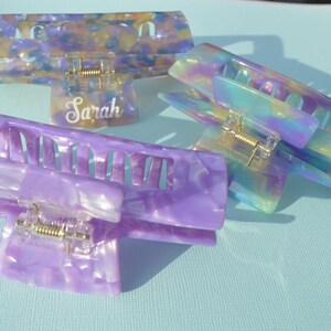 May include: Four iridescent hair claw clips in shades of purple, blue, and yellow. One clip has the name "Sarah" in white script. The clips have a rectangular shape with a gold-colored spring mechanism.