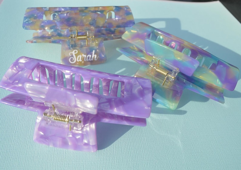 May include: Four iridescent hair claw clips in shades of purple, blue, and yellow. One clip has the name "Sarah" in white script. The clips have a rectangular shape with a gold-colored spring mechanism.