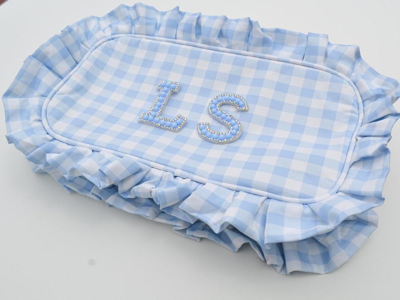 May include: A rectangular, light blue and white gingham patterned pillow with a ruffled edge. The pillow features the letters "LS" in a light blue, beaded font.