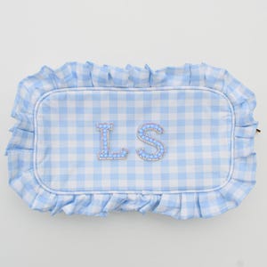 May include: A light blue and white gingham pattern pillow with a ruffled edge. The pillow features the embroidered letters "LS" in a sparkling, rhinestone-like design. The pillow is rectangular in shape.