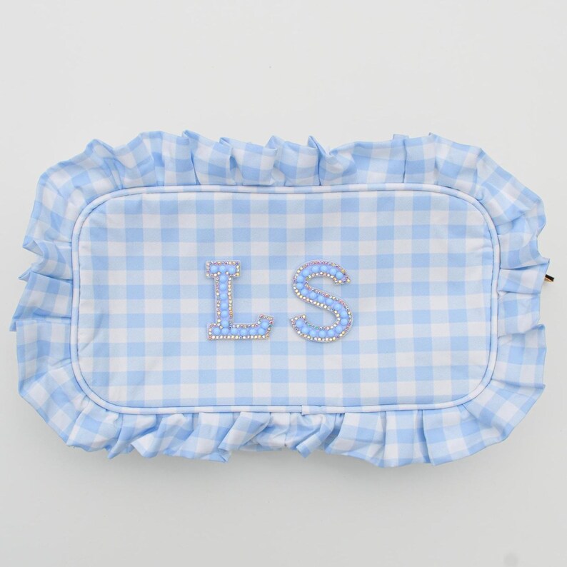 May include: A light blue and white gingham pattern pillow with a ruffled edge. The pillow features the embroidered letters "LS" in a sparkling, rhinestone-like design. The pillow is rectangular in shape.
