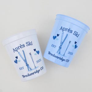 Puede incluir: Dos vasos de plástico, uno blanco y otro azul claro, con las palabras "Après Ski" y gráficos de esquí. Las tazas también presentan el texto "EST 2025" y "Breckenridge CO". Formas de corazón dispersas.