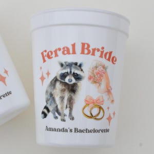May include: White plastic cup with the text "Feral Bride" in orange. The cup features a watercolor illustration of a raccoon, a bouquet of flowers, and two gold rings. The text "Amanda's Bachelorette" is also printed on the cup.