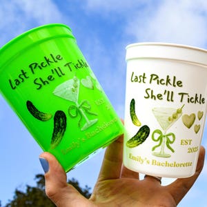 May include: Two plastic cups, one green and one white, are held up against a blue sky. The cups have the text "Last Pickle She'll Tickle" and a martini glass design with pickles and hearts. The green cup says "Emily's Bachelorette".