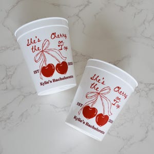 May include: Two white plastic cups featuring a red graphic of two cherries tied with a ribbon. The text reads "She's the Cherry on Top", "EST 2026", and "Kylie's Bachelorette" in red lettering. The cups have a tapered shape and a smooth finish.
