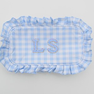 May include: A rectangular, light blue and white gingham pattern pillow with a ruffled edge. The pillow features the letters "LS" in a light blue, jeweled design.