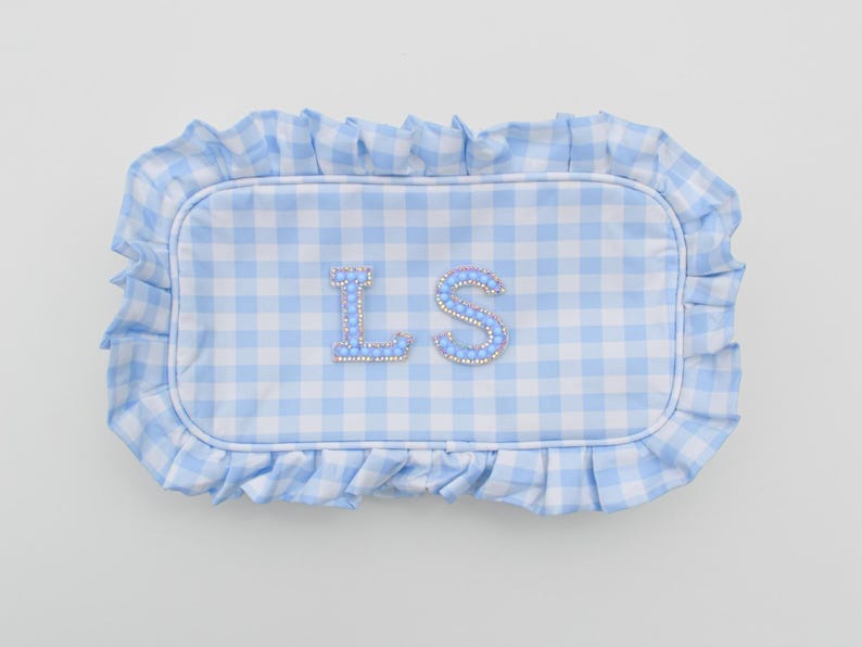 May include: A rectangular, light blue and white gingham pattern pillow with a ruffled edge. The pillow features the letters "LS" in a light blue, jeweled design.