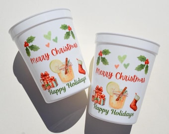 May include: Two white plastic cups with a festive Christmas design. Each cup features the text "Merry Christmas" and "Happy Holidays", along with illustrations of gifts, a drink, a stocking, holly, and hearts.