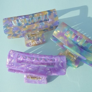 May include: Three colorful hair claw clips in shades of purple, yellow, and blue. One clip has the name "Sarah" printed on it. The clips are made of a marbled, translucent material and have a spring mechanism.