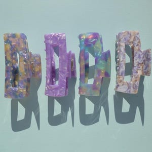 May include: Four colorful hair claw clips in shades of purple, yellow, and iridescent hues. Each clip has a unique marbled pattern and a sturdy clasp. The clips are arranged on a light blue surface, casting shadows.