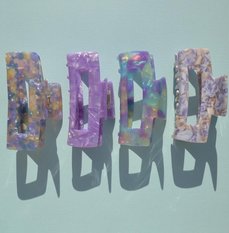 May include: Four colorful hair claw clips in shades of purple, yellow, and iridescent hues. Each clip has a unique marbled pattern and a sturdy clasp. The clips are arranged on a light blue surface, casting shadows.