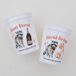 May include: Two white plastic cups with illustrations and text. One cup says "Just Feral" with a raccoon and champagne bottle. The other says "Feral Bride" with a raccoon, flowers, and rings. Both say "Amanda's Bachelorette".