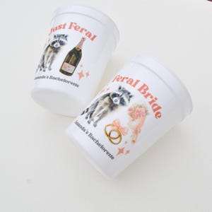 May include: Two white plastic cups with the text "Just Feral" and "Feral Bride" in coral. Each cup features a raccoon illustration, a champagne bottle, and wedding-themed graphics. The cups are labeled "Amanda's Bachelorette".