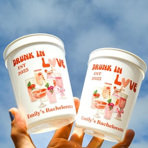 May include: Two white plastic cups with the text "DRUNK IN LOVE EST 2025" in orange script. The cups have a watercolor illustration of cocktails and the text "Emily's Bachelorette".