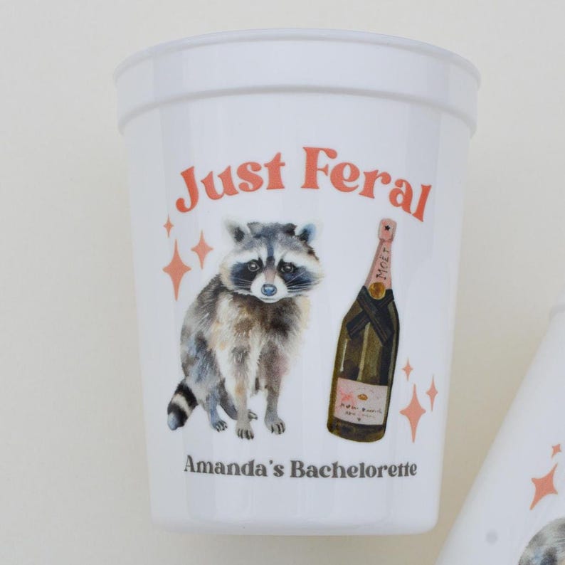 May include: White plastic cup with the text "Just Feral" in orange. The cup features a watercolor illustration of a raccoon and a champagne bottle. The text "Amanda's Bachelorette" is printed below the illustration.