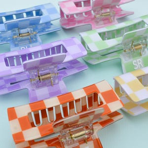 Puede incluir: Una colección de pinzas para el pelo a cuadros en tonos azul, rosa, verde, morado, naranja y amarillo. Los accesorios para el cabello tienen forma rectangular con mecanismo de resorte y están dispuestos sobre una superficie azul claro.