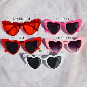 May include: Five pairs of heart-shaped sunglasses in different colours: double red, red, hot pink, light pink, and white. Each pair has the word "Bride" written on one lens.