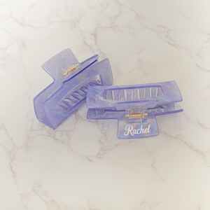 May include: Two translucent lavender hair claw clips with a pearlescent sheen. The clips have a rectangular shape with a gold-colored hinge and the name "Rachel" printed in white script on one clip. The clips are designed to hold hair in place.