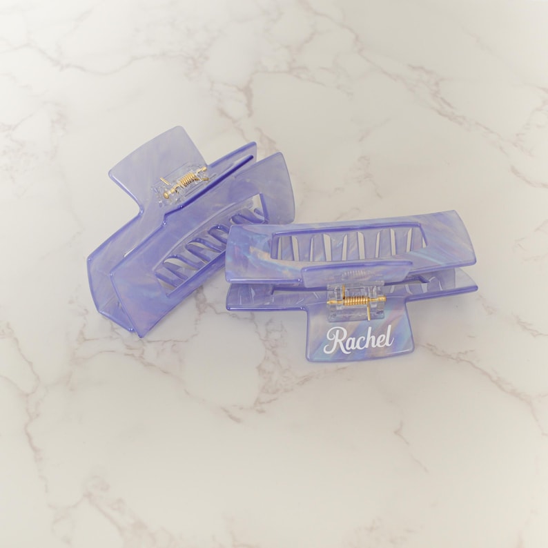 May include: Two translucent lavender hair claw clips with a pearlescent sheen. The clips have a rectangular shape with a gold-colored hinge and the name "Rachel" printed in white script on one clip. The clips are designed to hold hair in place.
