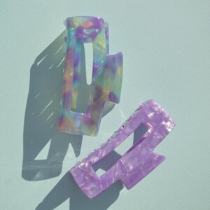 May include: Two rectangular hair claw clips. One clip is a mix of pastel colors, including purple, yellow, and blue. The other clip is a solid light purple color. Both clips have a geometric design and are on a light blue background.
