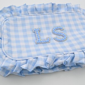 May include: A light blue and white gingham patterned pillow with a ruffled edge. The pillow features the letters "LS" embellished with light blue rhinestones. The pillow is rectangular in shape.
