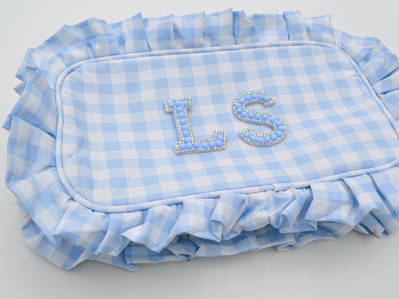 May include: A light blue and white gingham patterned pillow with a ruffled edge. The pillow features the letters "LS" embellished with light blue rhinestones. The pillow is rectangular in shape.