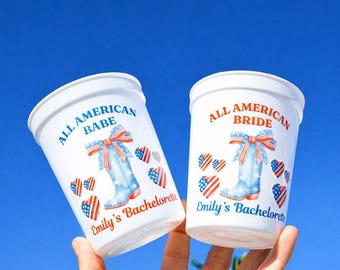 Könnte beinhalten: Zwei weiße Plastikbecher mit patriotischen Designs. Auf einem Becher steht "All American Babe" und auf dem anderen "All American Bride". Beide zeigen einen blauen Cowboy-Stiefel mit roter Schleife und Herzakzenten, mit dem Text "Emily's Bachelorette."