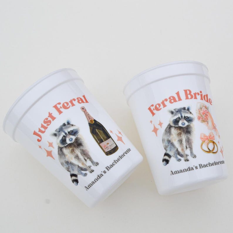 May include: Two white plastic cups with the phrases "Just Feral" and "Feral Bride" in red script. Each cup features a watercolor raccoon illustration and the text "Amanda's Bachelorette". One cup has a champagne bottle.