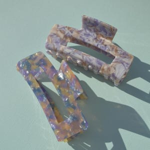 May include: Two marbled hair claw clips in shades of purple, yellow, and white. The clips have a geometric design with a rectangular shape and a claw-like grip. The clips are made of a glossy material.