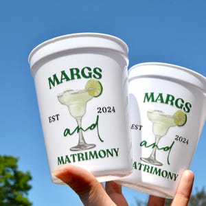Może przedstawiać: Dwa białe plastikowe kubki z zielonym tekstem i grafiką margarity. Tekst brzmi "Margs and Matrimony Est 2024".