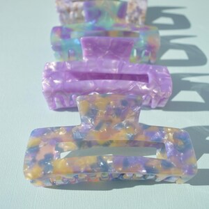 May include: Four rectangular hair claw clips in a row, each with a different marbled pattern. The colors include lavender, yellow, blue, and white. The clips are made of a shiny, hard plastic material.