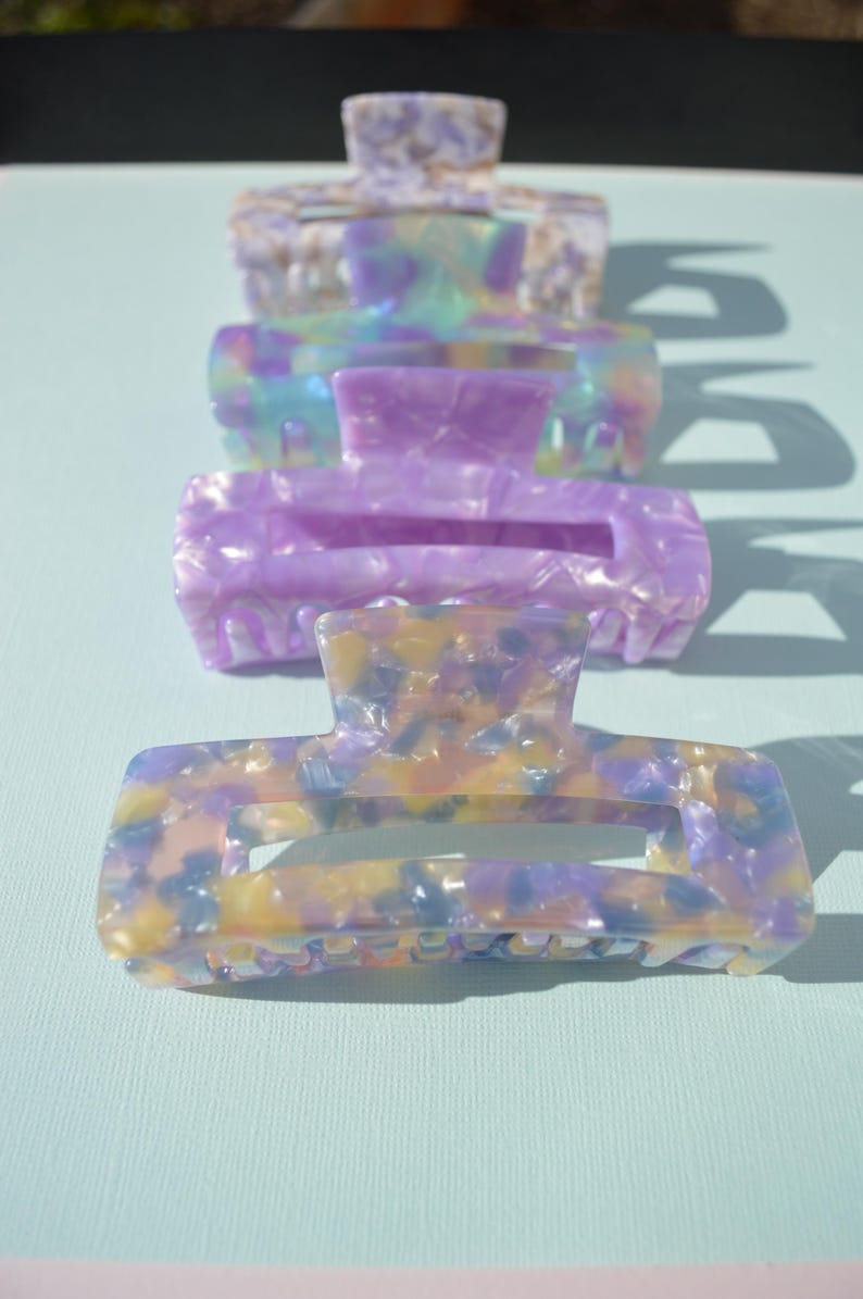 May include: Four rectangular hair claw clips in a row, each with a different marbled pattern. The colors include lavender, yellow, blue, and white. The clips are made of a shiny, hard plastic material.