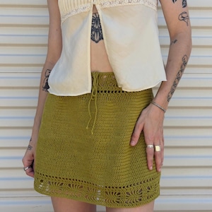 May include: A cream-colored, sleeveless top with a pleated front and lace trim is paired with a green crochet mini skirt. The skirt has a drawstring waist and a decorative, openwork hem. The outfit is casual and summery.
