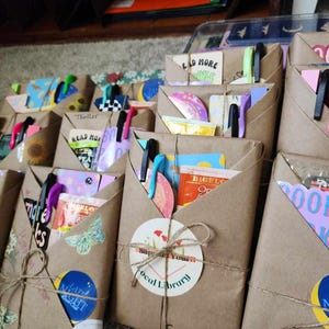 May include: A collection of brown paper packages wrapped with twine and decorated with colorful stickers, including a sticker that says "Support Your Local Library".