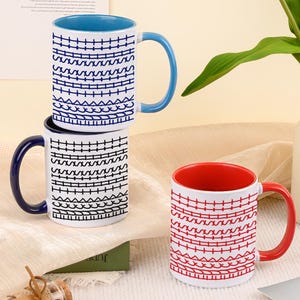 May include: Three ceramic mugs with patterned designs. One mug has a blue interior and handle, another has a red interior and handle, and the third has a dark blue handle. The mugs are stacked and arranged on a table.