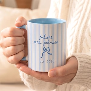 Puede incluir: Una taza de cerámica con un patrón de rayas verticales en azul claro y blanco. El texto "future mrs. johnson" está impreso en letra cursiva azul, seguido de un gráfico de un lazo azul y el texto "est. 2026" en una fuente azul sencilla.