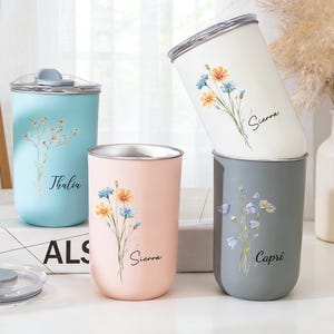 Puede incluir: Cuatro tazas de viaje de acero inoxidable en colores pastel: azul claro, rosa, blanco y gris. Cada taza presenta un diseño floral y un nombre en escritura cursiva. Las tazas tienen tapas plateadas.