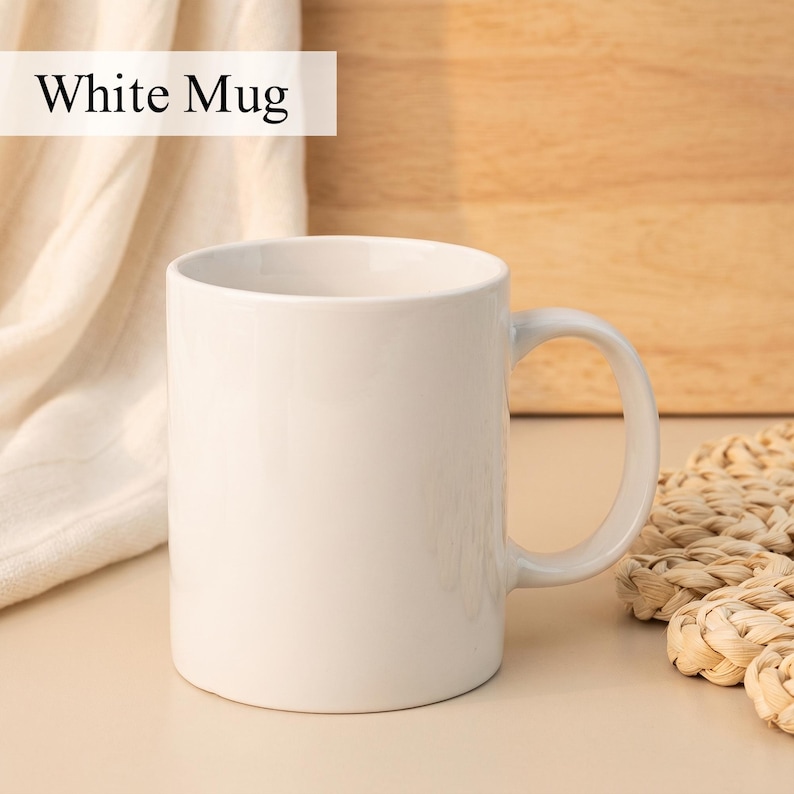 May include: A white ceramic mug with a curved handle. The mug is plain and has the text "White Mug" in a white rectangle at the top. The mug is sitting on a light-colored surface.