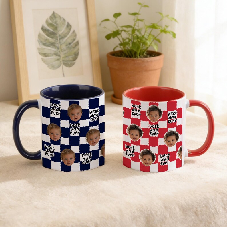 May include: Two ceramic mugs with the text "best dad ever" and a baby's face printed on them. One mug has a navy blue handle and interior with a white and navy blue checkered pattern. The other mug has a red handle and interior with a red and white checkered pattern.
