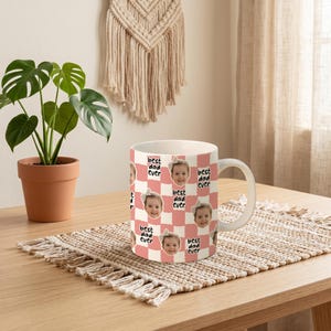 May include: A white ceramic mug with a pink and white checkered pattern. The mug features a repeating pattern of a child's face and the text "best dad ever". The mug is on a woven placemat on a wooden table.