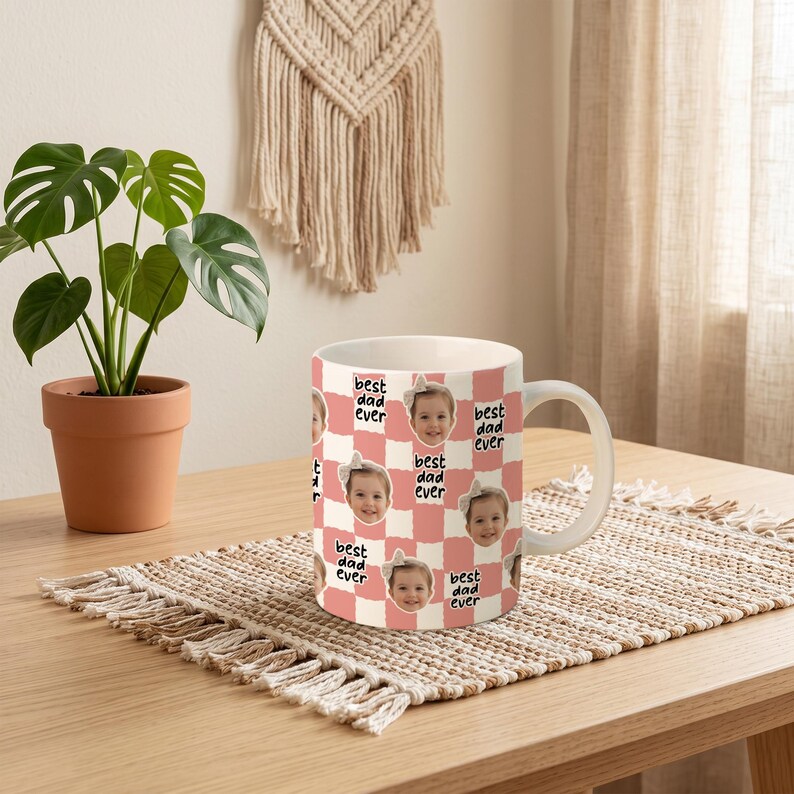 May include: A white ceramic mug with a pink and white checkered pattern. The mug features a repeating pattern of a child's face and the text "best dad ever". The mug is on a woven placemat on a wooden table.