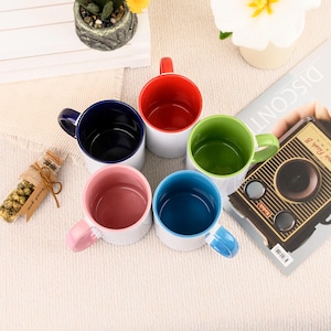 May include: A collection of five ceramic mugs with white exteriors and colorful interiors and handles. The interior colors are red, green, blue, pink, and navy. The mugs are arranged on a light-colored surface.