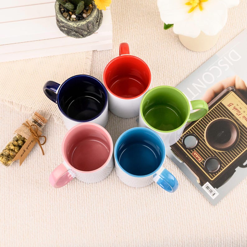 May include: A collection of five ceramic mugs with white exteriors and colorful interiors and handles. The interior colors are red, green, blue, pink, and navy. The mugs are arranged on a light-colored surface.