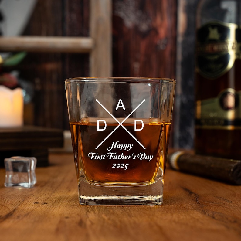 Op de afbeelding: Een vierkant whiskyglas gevuld met amberkleurige vloeistof. Het glas is ge&euml;tst met het woord "DAD" in een kruisontwerp en de tekst "Happy First Father's Day 2025". Het glas staat op een houten oppervlak.