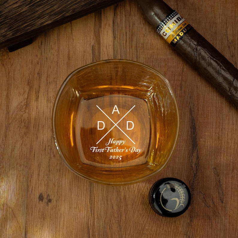 Op de afbeelding: Een vierkant whiskyglas met het woord "DAD" en de tekst "Happy First Father's Day 2025" op de bodem gegraveerd. Een sigaar en een zwarte flesstopper liggen naast het glas op een houten oppervlak.